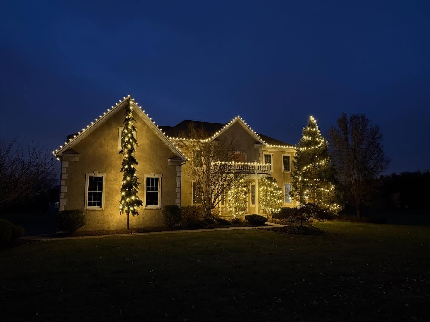 Warm white Christmas lights on the roofline, trees, and shrubs of a stucco home at dusk